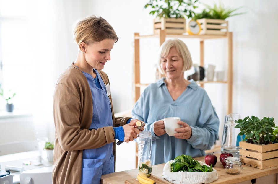 a caregiver offers meal prep and cooking services for an elderly client in her home, while the woman looks on happily with a cup of tea